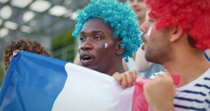 Group of multi-ethnic colleagues shouting and cheering for France in big tournament. Shaking with their hands French flag and smiling. Wearing wigs and small painted French flags on their faces.