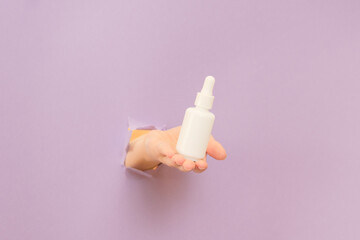 Hand holding glass bottle with dropper lid. White container with cosmetic product, serum (essential oil ) on lilac background. Concept of beauty