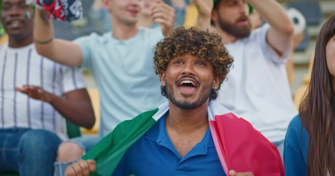 Handsome man holding Italian flag during sport tournament. Young people actively cheering their team. Singing song or shouting words of support. Happy fans having fun during championship.
