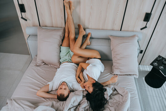Playful couple lying upside down on bed, intertwined legs raised against a wooden wall, creating a relaxed, fun atmosphere