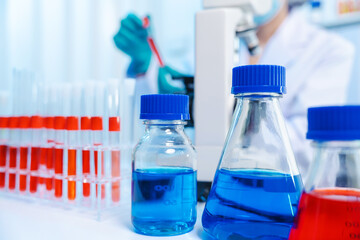 Asian people scientist in lab coat and protective gloves working with test tubes with green and red liquids, with microscope and other test tubes in the background in laboratory.