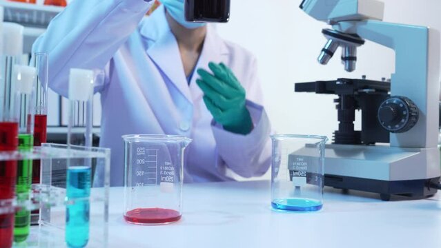 Asian people scientist in lab coat and protective gloves working with test tubes with green and red liquids, with microscope and other test tubes in the background in laboratory.