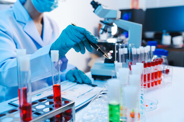 Asian people scientist in lab coat and protective gloves working with test tubes with green and red liquids, with microscope and other test tubes in the background in laboratory.