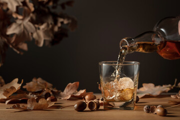 Whiskey with ice on a wooden table with dried-up oak leaves.