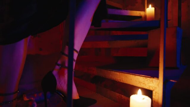 following shot of Model's legs in high heels ascend stairs, stepping over a red rose. On the stairs: a pumpkin, book, and candles. Black dress, nighttime indoors