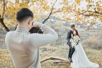 Professional wedding photographer taking pictures of the bride and groom in nature in autumn - obrazy, fototapety, plakaty