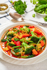 vegetable soup with green beans, broccoli, carrot, celery, spinach, cabbage, zucchini, tomatoes in white bowl on white wooden table with ingredients and spoon at background, vertical view, close-up