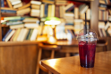 A red lemon cocktail with ice cubes and a straw stands on a wooden table in the library. Large bookshelves, table, chair and lamp on the background at the cafe. Break while studying.