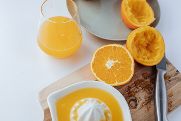 There is a manual juicer on a wooden cutting board, next to it is a knife and half a squeezed orange. a glass of juice and a plate of oranges. White background