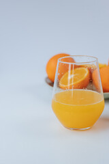a glass glass with orange juice in the foreground, a plate with oranges in the background. White background
