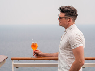 Stylish man with a glass of drink standing on the empty deck of a cruise liner against the backdrop...