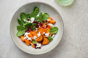 Plate with roasted sweet potato, feta cheese, baby spinach and pomegranate, above view on a beige stone background, horizontal shot