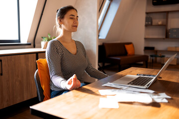 Brunette woman freelancer sitting in lotus pose and meditating with hands in mudra gesture on chair