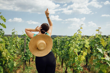 Young woman in black pants and wide brimmed straw hat stands in graceful pose and looks at vineyard.