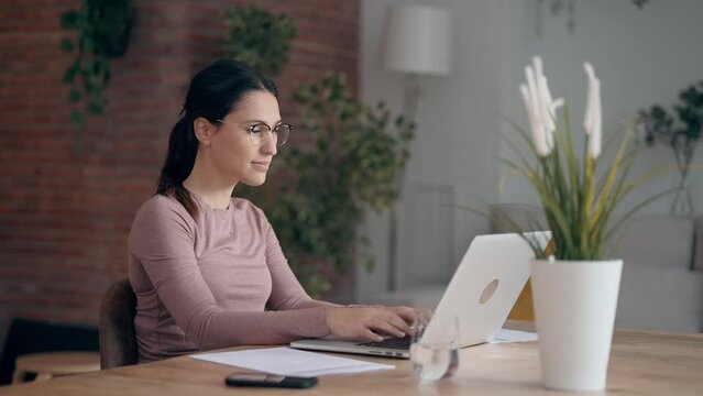 Video of concentrated beautiful business woman working with laptop while drinking coffee in living room at home.
