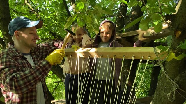 Family bonding. The joy of childhood by spending leisure time together in the great outdoors. Dad and elementary age kids joyfully build a summer treehouse, creating delightful childhood memories.