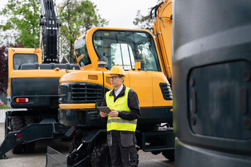 Engineer in a helmet with a digital tablet stands next to construction excavators.