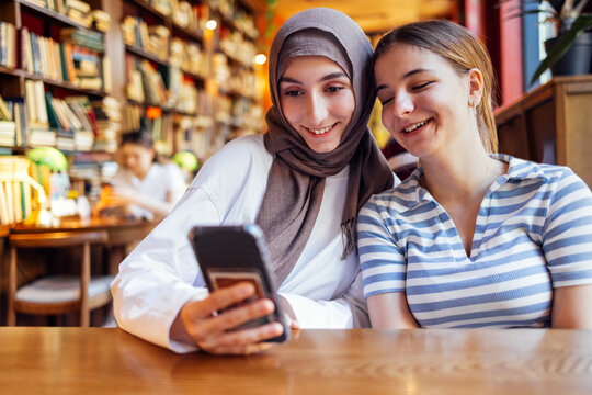 Two teenage girls of different nationalities and religions are sitting in library and looking at phone. Charming female students have rest from classes and use mobile gadget. Bookshelves on background - Powered by Adobe