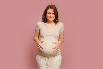 A pregnant woman holds onto a sore stomach on a studio pink background. Pregnancy in a woman with a belly, copy space