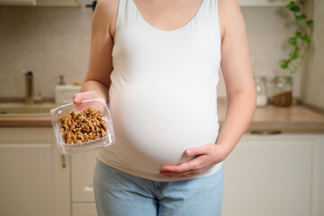 A pregnant woman holds walnuts nuts in her hands while standing in her home kitchen. Pregnancy and...
