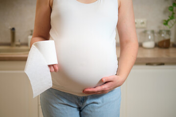 A pregnant woman holds toilet paper in her hands while standing in her home kitchen. Pregnancy and...