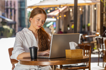 Fototapeta premium Redhead child girl working study education online distant job with laptop in city sunny street cafe restaurant browsing website chatting outdoors during break. Smiling woman tourist freelancer