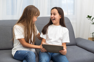 Cute little girl and older sister playing together smiling and having bonding time using a laptop on couch at home. Happy family Siblings relationship and digital technology lifestyle concept.