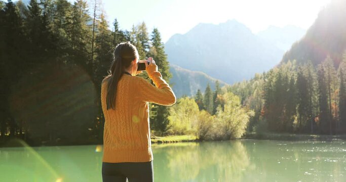 Young woman photographing at an alpine lake in beautiful autumn scenery. Female photographer with camera at a scenic lake on sunny autumn day. Europe travel concept.