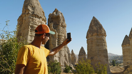 A man in a yellow T-shirt and baseball cap takes a picture of the mountains with his phone. Love Valley in Cappadocia. A blogger shoots a story on a smartphone about cliffs in Turkey.