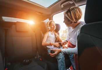 Inside the car photo of a mother fastening with safety auto belt her little daughter girl sitting in child seat. Girl listening music using headphones. Family values, traveling and technology concept - obrazy, fototapety, plakaty