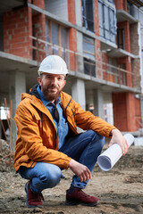Cheerful man in protective work helmet holding architectural building plan and smiling while...