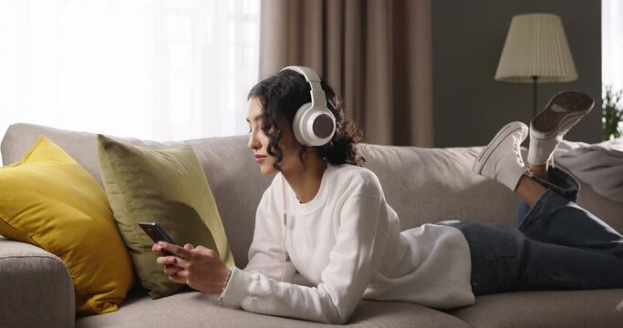 Young girl lies on sofa and listens to music on headphones large panoramic window lets lot of light into room. Music fills with joy, creating delightful mood. With headphones on girl rocks to music.