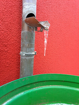 Metallenes Fallrohr der Regenrinne vor roter Wand. Ein Eiszapfen h&auml;ngt am Auslauf des Rohrs, direkt &uuml;ber einer gr&uuml;nen Regentonne. Klare Formen,  harmonische Farben: Rot, Gr&uuml;n und metallisch Graubraun.
