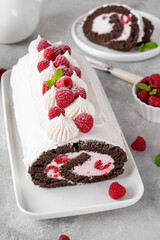 Chocolate roll cake with cream and fresh raspberries on a white plate on a gray background. Delicious holiday dessert.