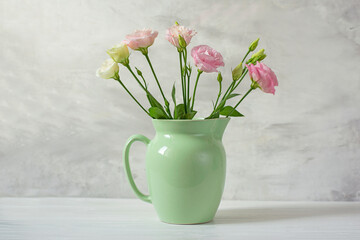 A bouquet of delicate pink and white eustoma lisianthus flowers in a light green ceramic jug on a light gray wall background. 