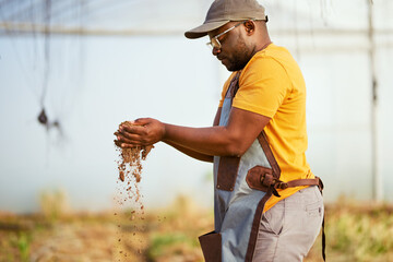 Black farmer lets soil fall through his hands, greenhouse in background. 