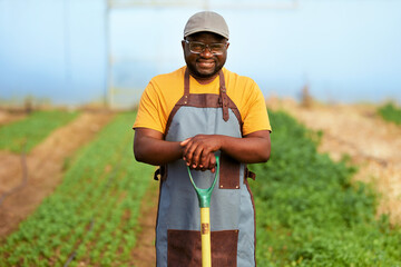 Black farmer standing in greenhouse with spade, crops in background, smiling.