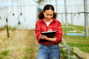 Multi-ethnic female farmer in greenhouse, writing notes, crops in background
