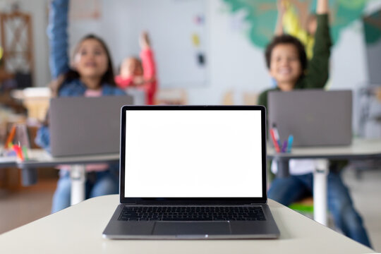 Laptop computer with blank mockup screen on teachers table with junior children pupils raising hands in classroom. Education software website technology ads concept