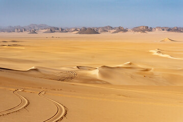 Landscape of Erg Admer in the Sahara desert, Algeria. The golden sand of Erg Admer with, in the distance, the rock formations of Tassilis. In the foreground, we can see the tracks left by jeep tires - 704499219