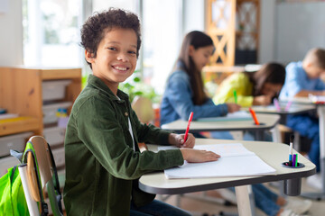 Smiling black boy writing in classroom, learning, knowledge and study. Latin male pupil at school,...