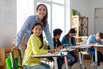 Cheerful teacher working with elementary school girl at desk. Happy teacher and girl smiling at camera in classroom