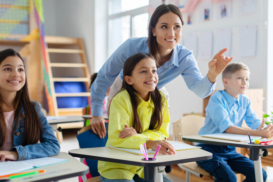Cheerful teacher stands among engaged pupils, pointing at blackboard while explaining, creating an interactive and vibrant learning atmosphere