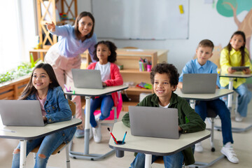 School children using laptops, female teacher pointing at blackboard during exam test at computer...