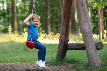 Smiling blond boy on red swing on green sunny forest background. Horizontal portrait , there is free space for text