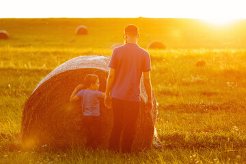 A father with his son near a large bale of straw, shot from the back in front of the golden evening...