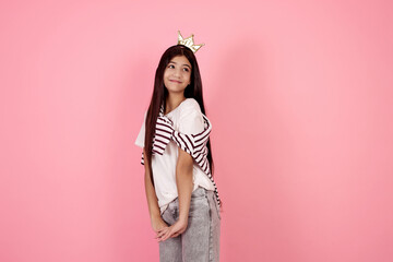 Young smiling and shy teenage girl with crown on pink background in studio.