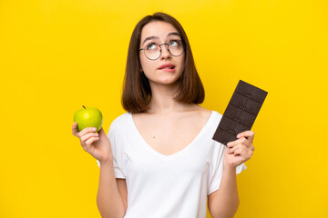 Young Ukrainian woman isolated on yellow background having doubts while taking a chocolate tablet...