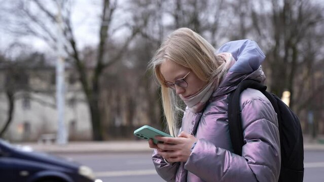 Schoolgirl stands outside and uses her phone. Teen girl with backpack waits for a bus after school. Young blond hair with glasses girl portrait.
