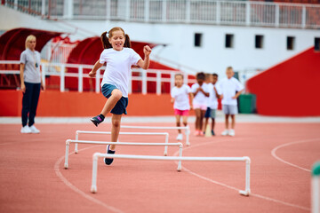 Happy girl jumping over obstacles while running at stadium.